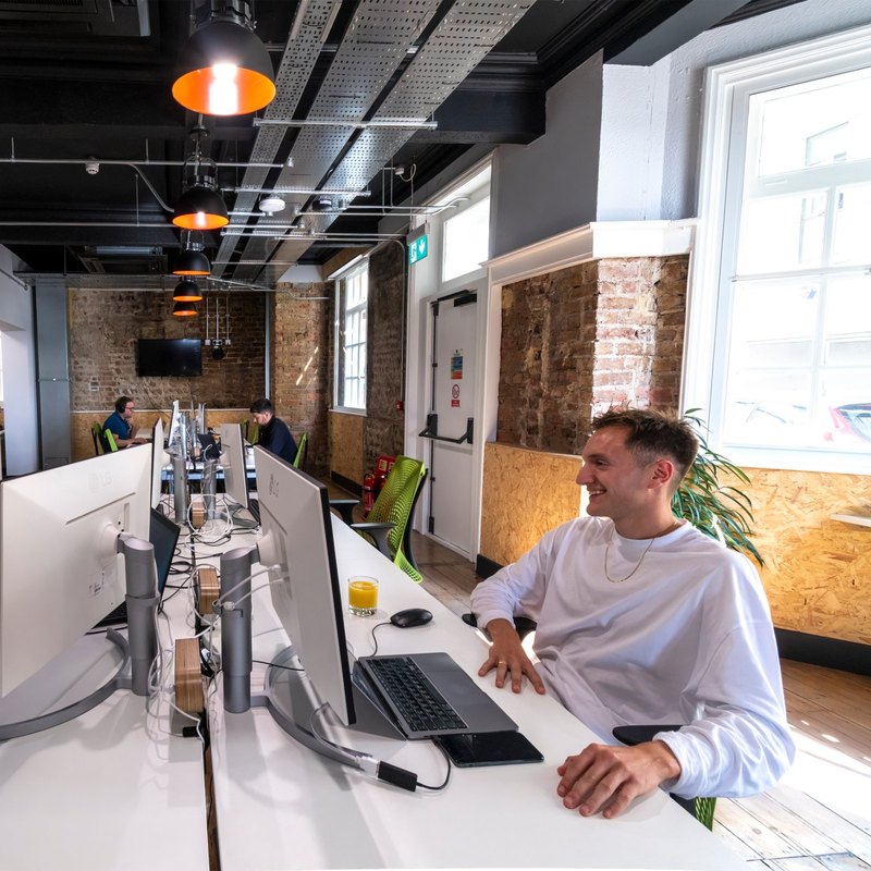 An FMG colleague smiling at his desk in the Clarendon Mansions office, with exposed brick, industrial lighting and bright windows