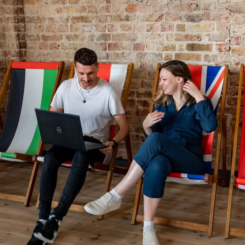 Two FMG colleagues chatting on flag deckchairs against an exposed brick wall at Clarendon Mansions